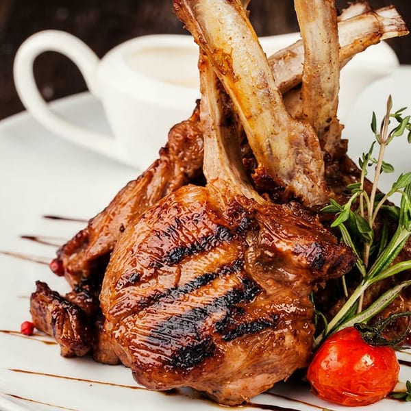 Grilled lamb chops with rosemary, a cherry tomato, and garlic on a white plate.
