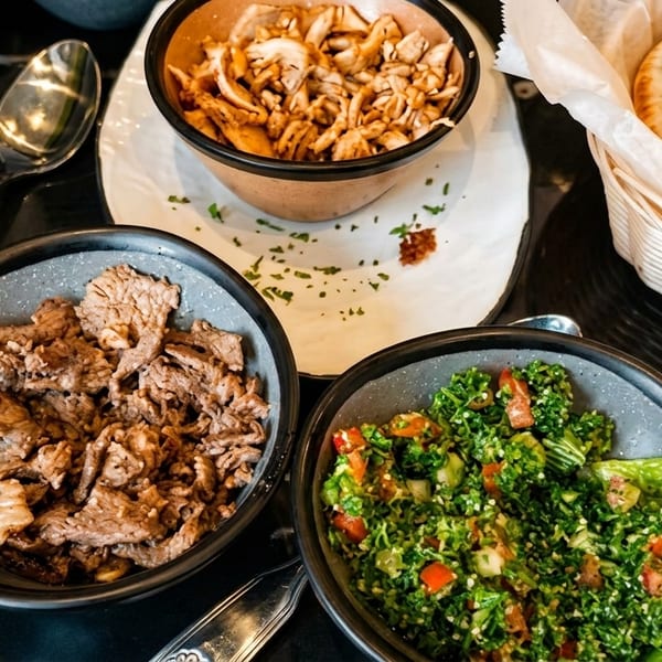 Bowls of beef, chicken, tabbouleh salad, and a basket of pita bread on a dark table.