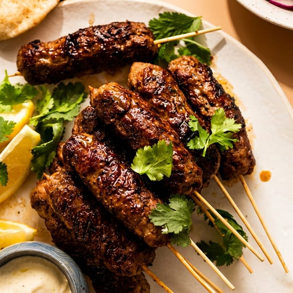 Grilled kofta kebabs with lemon wedges, cilantro, pita bread, and dipping sauce on a white plate.