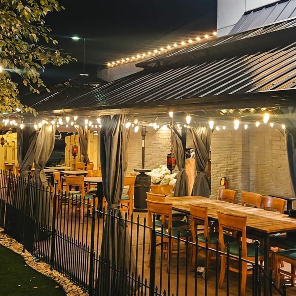 Outdoor restaurant patio at night with string lights, covered seating areas, and a tree wrapped in lights.